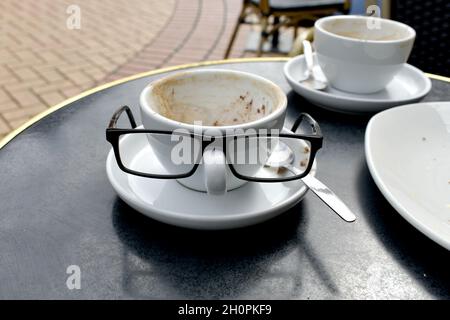 Nahaufnahme von weißen Kaffeetassen auf einem Tisch. Seine und ihre Tassen mit einer Mans-Brille, die auf dem Griff thront, sehen aus wie ein lustiges Gesicht vor einem Café-Restaurant Stockfoto