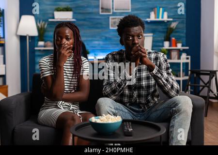 afroamerikanische Menschen sehen sich im Fernsehen einen Theaterfilm an und werden im Wohnzimmer geschockt. Schwarze Frau und Mann mit der Hand über den Mund auf den Fernseher, während sie auf die Kamera schauen. Afro-Paar Stockfoto