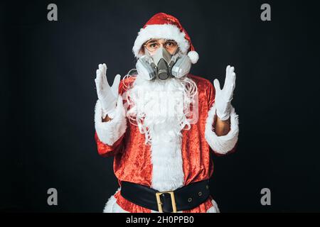 Mann, der als Weihnachtsmann gekleidet ist und eine Kohlefilter-Maske trägt, um einen Virus zu vermeiden, auf schwarzem Hintergrund. Weihnachtskonzept, Weihnachtsmann, Geschenke, Celebra Stockfoto