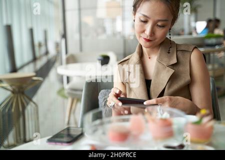 Reife asiatische Frau, die im modernen Restaurant beim Nachmittagstee auf das Handy schaut Stockfoto