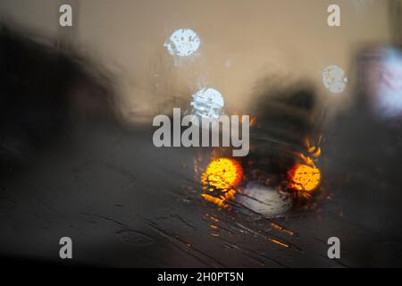Regnerisches Wetter, Regentropfen durch das Glas, abstrakter Hintergrund vor dem Licht von Autos und Ampeln, defokussiert Stockfoto