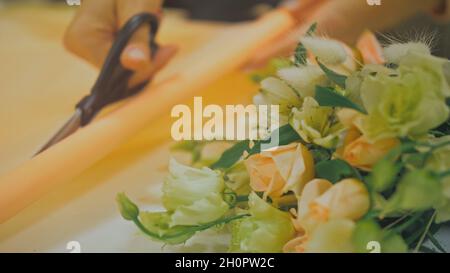 Weibliche Floristin macht Blumenstrauß, sie schneidet Verpackungspapier in Blumenladen zu arbeiten. Cute junge professionelle macht Blumenstrauß im Blumengeschäft. Stockfoto