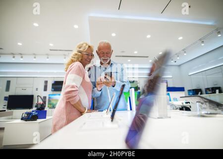 Glückliches Seniorenpaar auf der Suche nach einem neuen Smartphone. Sie stehen in der Nähe stehen im Technikgeschäft. Mann hält Smartphone, während Frau schaut. Stockfoto
