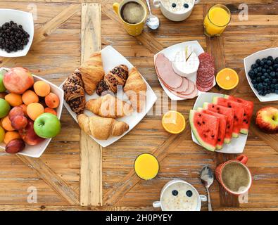 Morgens gibt es ein gesundes Frühstück auf einem Holztisch, eine Auswahl an verschiedenen leckeren Speisen, Haferflocken mit Milch, frisch gebackene Croissants und Obst Stockfoto