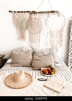 Modernes schlafzimmer im scandi Boho-Stil mit dekorativen Kissen, Korbhut, Früchten und offenem Buch auf dem Bett, Makrame-Dekorationen und Traumfänger-Hangi Stockfoto