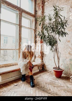 Sepia-Stil launisch Foto von nachdenklichen jungen Frau in langen Kleid mit Vintage-Kamera in der Hand sitzt auf Fensterbank in alten ungepflegten Zimmer mit eingetopften grünen p Stockfoto