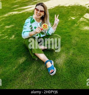 Fröhliche, fröhliche junge Frau, die mit der Seifenblasenmaschine Blasen bläst und lacht, während sie auf grünem Gras im Park sitzt und sich auf dem sonnigen Summ amüsiert Stockfoto