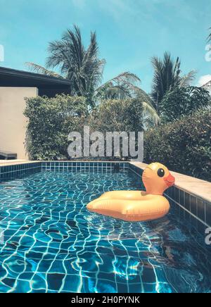 Aufblasbarer Ring aus gelber Ente, der am heißen, sonnigen Sommertag im Swimmingpool mit Palmen im Hinterhof eines modernen Ferienhauses oder Luxushotels schwimmt Stockfoto