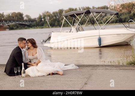 Konzept des glücklichen Familienlebens, Braut und Bräutigam in Hochzeit weißes Kleid auf Betonufer des Stadtflusses im Hintergrund ist weißes Motorboot, verwendet forwa Stockfoto