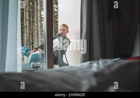 Ein Mann mittleren Alters in offener, warmer Strickjacke und Jeans sitzt auf einem Waldhausbalkon und genießt die frische herbstliche kalte Luft. Alltagslebheit Stockfoto