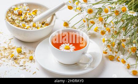 Weiße Tasse heißer Kamillentee, ein Mörser mit trockenen Gänseblümchen-Knospen und ein Bouquet von Matricaria-Kamilla auf betontem Hintergrund. Kräutergetränk, gesundes li Stockfoto