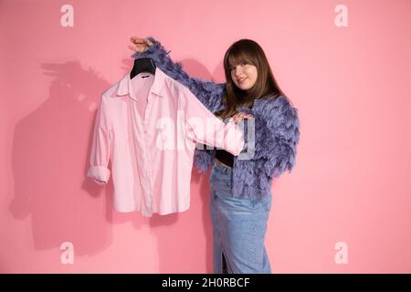 Junge Frau Fashion Stylistin hält Kleiderbügel mit Hemd auf rosa Hintergrund im Studio. Einfaches Garderobenkonzept. Stockfoto