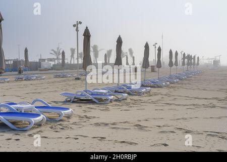 Nebliger Strand mit verschwommenen Reihen von geschlossenen Sonnenschirmen, Sonnenliege in der Nebensaison an nebligen Tagen. Dichter Nebel am Strandbereich, leere Sonnenliegen, geschlossene Sonnenschirme. Stockfoto