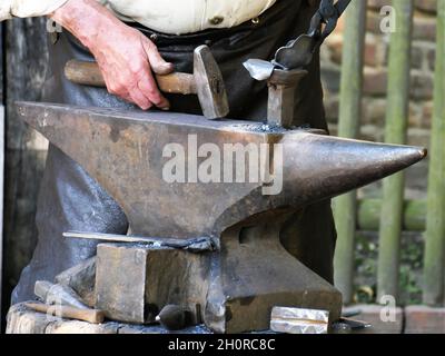 Stahl Amboss, auf dem ein heißes Werkstück liegt, die Hand des Schmieds verarbeitet ein Werkstück mit Hilfe eines Hammers und einer Zange , fuzzy backgr Stockfoto