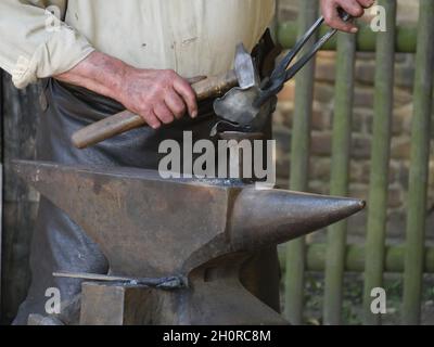 Stahlanvil, auf dem ein heißes Werkstück liegt, bearbeitet die Hand des Schmieds ein Werkstück mit Hilfe eines Hammers und einer Zange, des Hammers Stockfoto