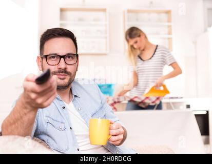 Junger, hübscher Mann, der auf dem Sofa sitzt und die Fernbedienung des fernsehers hält, während seine Frau im Hintergrund bügelt und Aufgaben erledigt Stockfoto