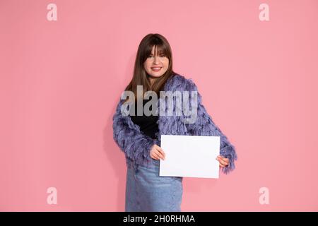 Foto von niedlichen jungen dunkelhaarigen Frau. Hält leere, saubere, weiße Blatt Papier für Inschrift lächelt, schaut in den Rahmen. Foto auf rosa Hintergrund in Stockfoto