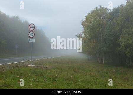 Gefährlicher Nebel auf der Straße in den Bergen, Tempolimit-Straßenschild Stockfoto