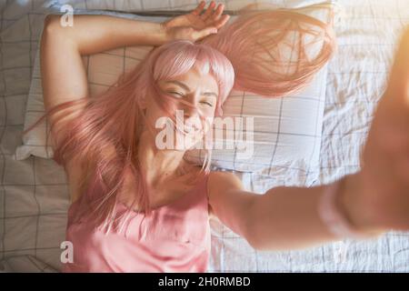 Fröhliche Dame mit leuchtend rosa Haaren nimmt Selfie auf einem bequemen Bett liegend Stockfoto