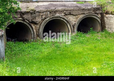 Große Abflussrohre oder Abwässer leiten Abwasser mit grünem Gras und Abfall in den Abwasserkanal. Stockfoto