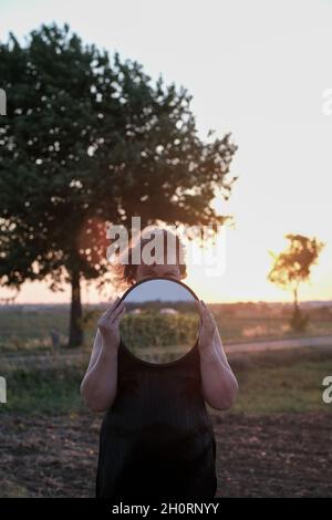 Porträt einer Frau, die auf einem Feld steht und einen Spiegel vor ihrem Gesicht hält, Frankreich Stockfoto