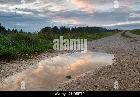 Spiegelungen eines Sonnenuntergangs Himmel in einer regen Pfütze auf einer Schotterstraße in ländlicher Landschaft. Gesehen in Deutschland im Oktober Stockfoto