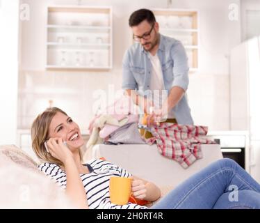 Schöne junge Frau, die sich auf dem Sofa ausruhte und auf dem Handy sprach, während der Mann ihr Hemd im Hintergrund bügelt. Ehemann macht Aufgaben Konzept Stockfoto