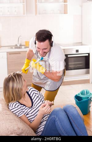 Schöne junge Frau ruht sich aus, während der Mann den Boden wischt und singt. Ehemann macht Aufgaben Stockfoto