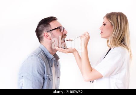 Junge Ärztin füttert männlichen Patienten mit einem Löffel voller verschiedener Medikamente Stockfoto