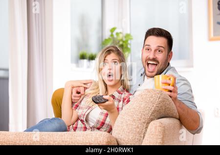 Junges glückliches Paar, das auf dem Sofa sitzt und Fernsehen genießt. Angeregter Ausdruck auf Gesichtern Stockfoto