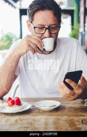 Schöner erwachsener Mann, der Kaffee trinkt und auf dem Handy nach E-Mails oder Niftigungen überprüft. Moderne, reife Leute im Café genießen Wireless Internet Stockfoto