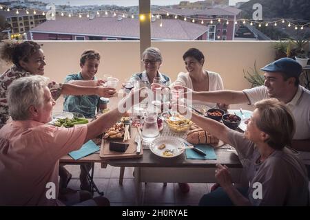 Eine Gruppe von Freunden gemischter Altersgruppen, die abends auf der Terrasse mit Blick auf die Stadt zu Hause mit Gläsern toasten - gemischte Generationen haben Spaß Stockfoto