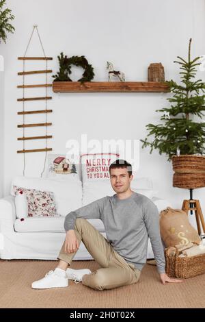 Hübscher junger Mann in legerem Outfit sitzt auf dem Boden im Wohnzimmer, das zu Weihnachten geschmückt ist Stockfoto