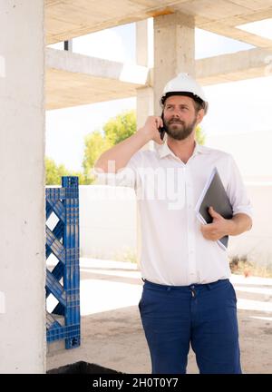 Zuversichtlich, Architekt. Hübscher junger Mann Bauarbeiterhelm holding Blaupause und sprechen auf dem Handy stehen im Freien und gegen den Bau stru Stockfoto
