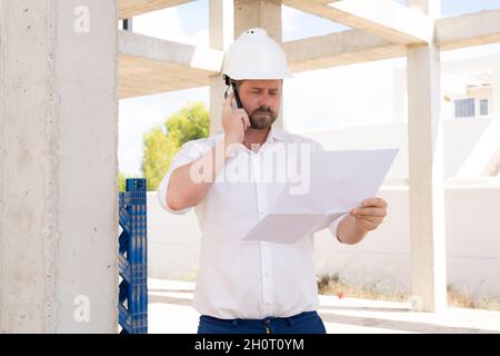 Zuversichtlich, Architekt. Hübscher junger Mann Bauarbeiterhelm holding Blaupause und sprechen auf dem Handy stehen im Freien und gegen den Bau stru Stockfoto