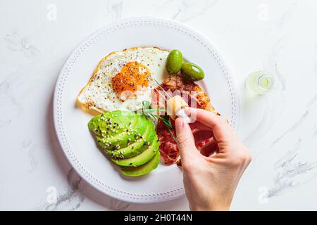 Ätherisches Cannabisöl tropft in die Nahrung. Keto Frühstückskonzept. Spiegelei mit Avocado und gebratenem Speck auf einem weißen Teller. Stockfoto