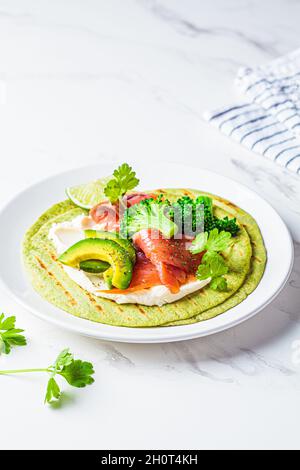 Grüne Tortilla mit Brokkoli, Avocado, Frischkäse und gesalzenem Lachs auf weißem Marmorboden. Vegetarisches Fast Food. Stockfoto