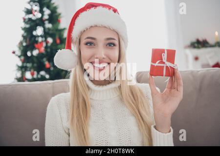 Foto Porträt lächelnde Frau sitzt auf dem Sofa trägt weißen gestrickten Pullover Kopfbedeckung halten kleine Geschenk-Box Stockfoto