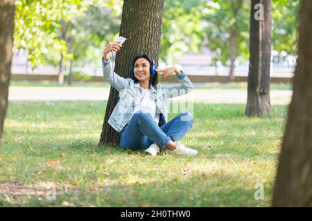 Junge asiatische Frau mit blauen Kopfhörern nimmt ein Selfie auf oder streamt online, während sie unter dem Baum im Stadtpark sitzt. Modernes Technologiekonzept. Stockfoto