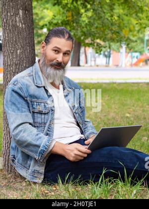 Foto eines bärtigen Mannes, der mit einem Laptop online arbeitet. Er schaute auf die Kamera. Vertrisches Foto. Stockfoto