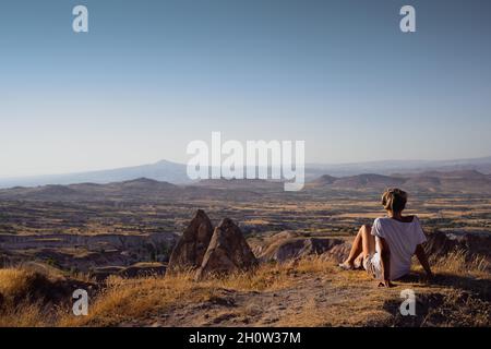 Rückansicht einer blonden kaukasischen jungen Frau mit weißem Hemd und Shorts, die auf einem Hügel sitzt und die wunderschöne Tallandschaft von Cappadocia Turk bewundert Stockfoto