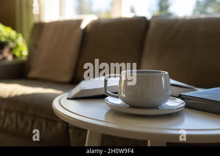 Kaffee auf einer Untertasse auf einem runden weißen Couchtisch mit offenem Buch, bibel, vor einer Ledercouch Stockfoto