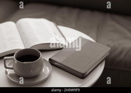 Kaffee auf einer Untertasse auf einem runden weißen Couchtisch mit offenem Buch, bibel, vor einer Ledercouch Stockfoto