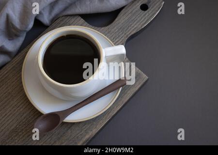 Einfache Tasse Kaffee auf einer Untertasse mit einem kleinen Holzlöffel und grauem Leinen auf schwarzem Hintergrund mit Kopierraum Stockfoto