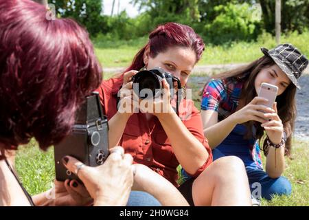 Verschiedene Generationen von lateinischen Frauen im Freien, jede von ihnen hält eine Kamera, die zu ihrer Generation passt: Analoge, digitale und mobile Technologie, Stockfoto