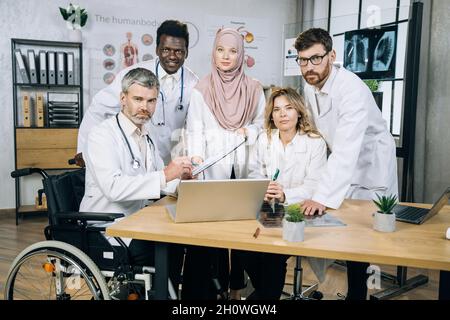 Eine Gruppe von fünf verschiedenen Medizinern mit einem männlichen Anführer, der im Rollstuhl sitzt und während der Konferenz im Büro lächelt. Einbeziehung von Menschen im Rollstuhl. Stockfoto