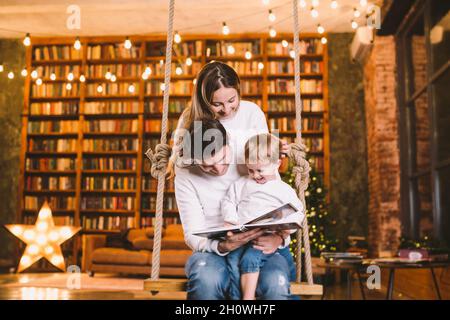 Familie mit Baby Entspannung zu Hause Schaukel Sitz in gemütlichen dunklen Wohnzimmer am Heiligabend. Die Familie hat Spaß auf der Wippe. Winterabend mit Familie und KI Stockfoto