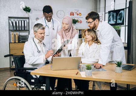 Reifer Arzt im Rollstuhl Unterschrift Papier auf Zwischenablage, die Frau in Hijab und Laborkittel halten. Gruppe von multiethnischen Mitarbeitern, die in der Nähe stehen und auf einem Laptop suchen. Stockfoto