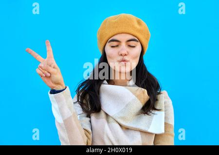 Foto von schönen verträumt Dame Schlag Luft Kuss zeigen V-Zeichen schließen Augen tragen beige Baskenmütze und Mantel isoliert Biue Farbe Hintergrund Stockfoto