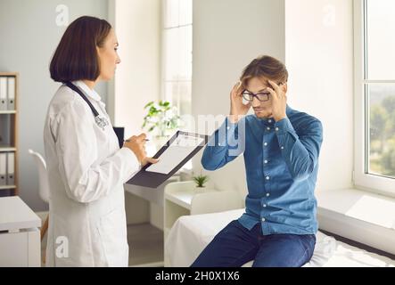 Ein männlicher Patient im Krankenhaus beschwert sich beim Arzt über Kopfschmerzen, indem er seine Finger an seine Schläfen hält. Stockfoto